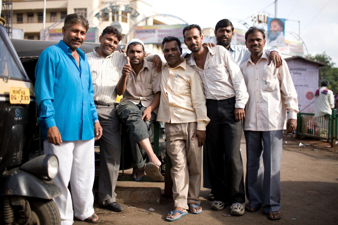 Seven men in casual and formal wear stand together smiling in front of a vehicle in Pune, India.