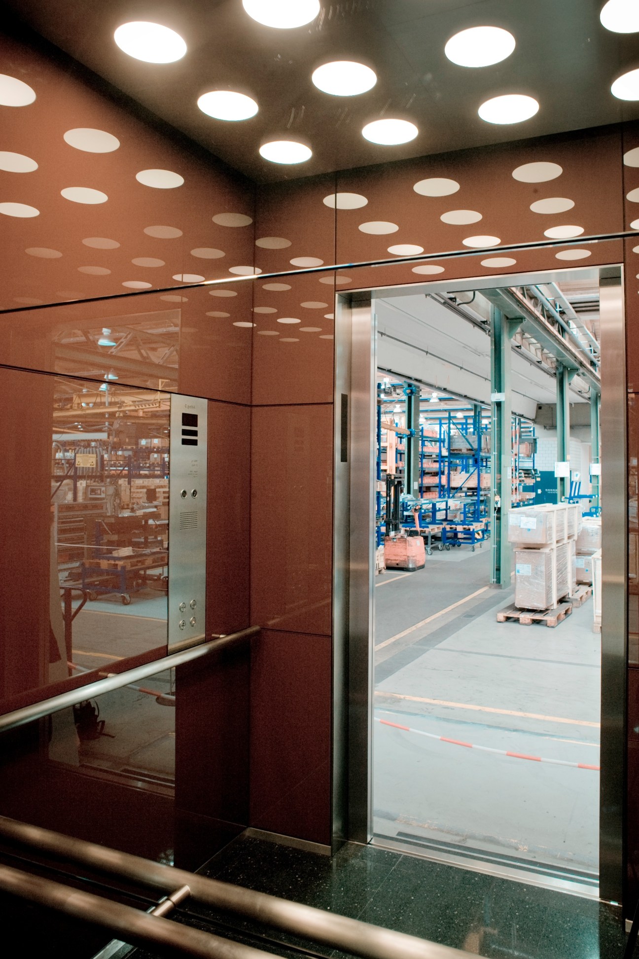 Modern elevator interior with terracotta walls, recessed ceiling lights, and glass doors opening to a factory floor.