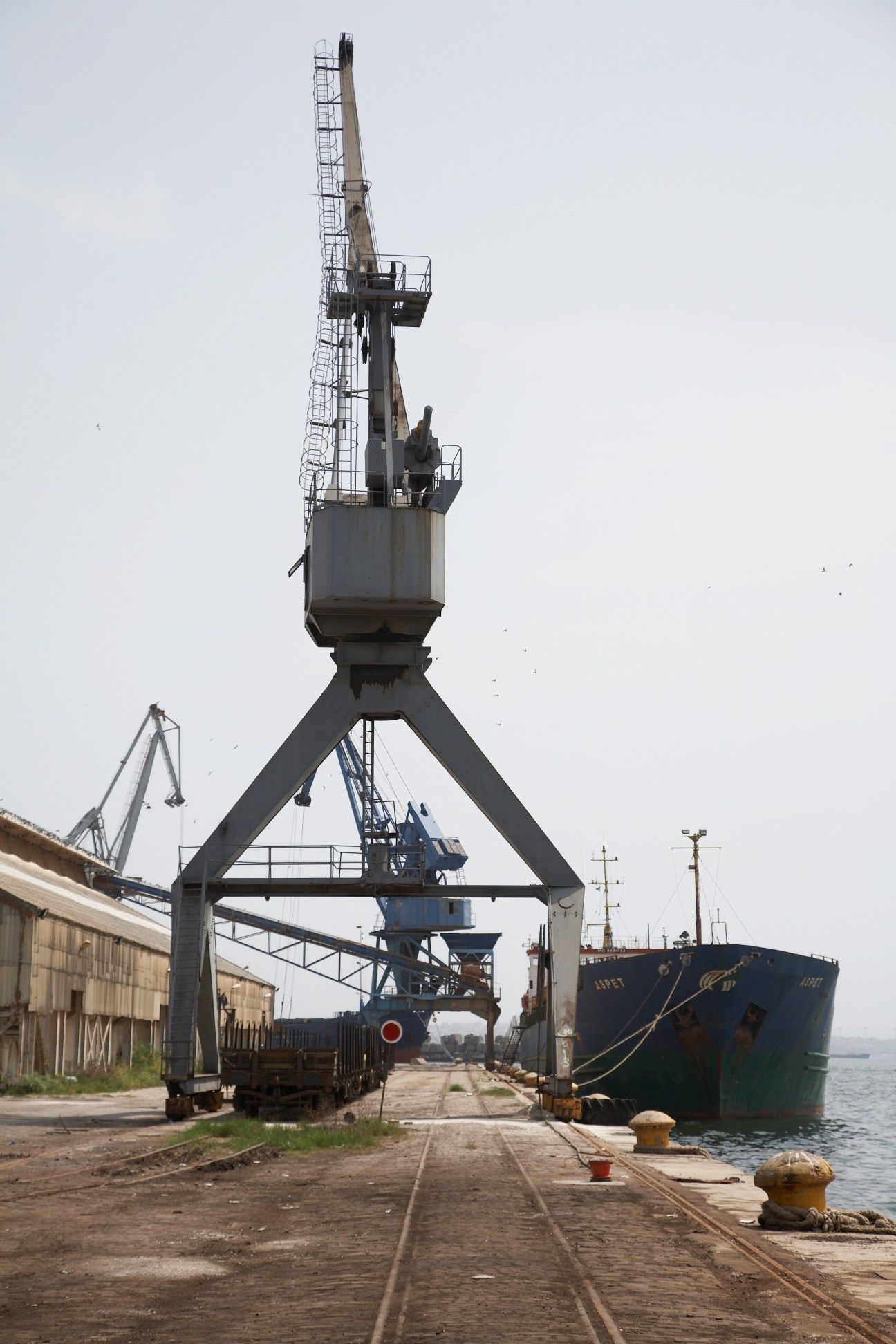 Industrial container crane towering over railway tracks and moored cargo ship in Thessaloniki harbour.