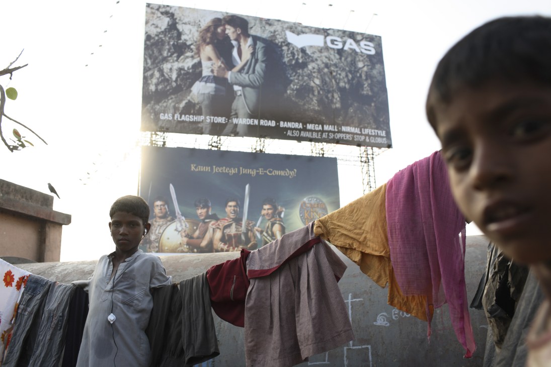Person hanging laundry in slum area with large commercial billboards advertising luxury brands in background.