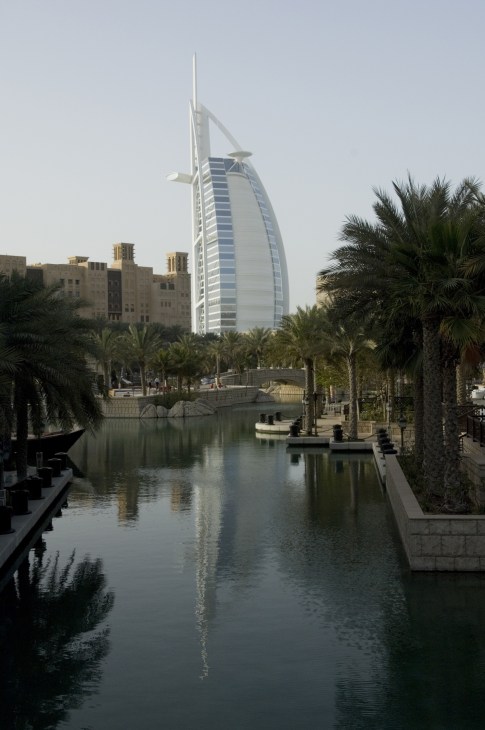 The Burj Al Arab hotel in Dubai with its distinctive sail-shaped white architecture reflected in a waterway lined with palm trees.