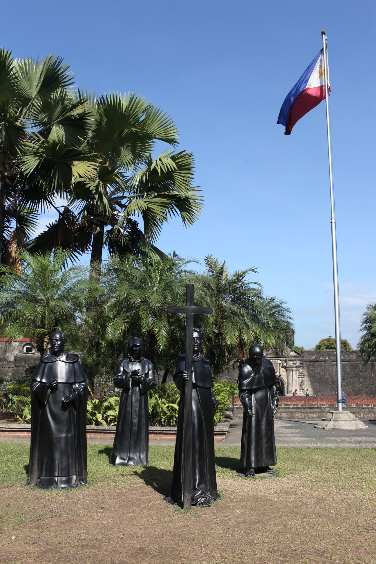 Bronze statues of religious figures with a cross in Intramuros, Manila's historic walled district, with Philippine flag flying nearby.