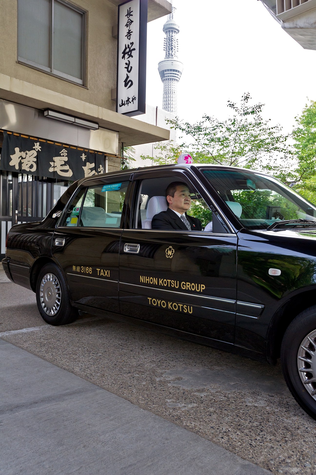 Black Nihon Kotsu taxi with driver in formal attire parked on street with Tokyo Skytree visible.
