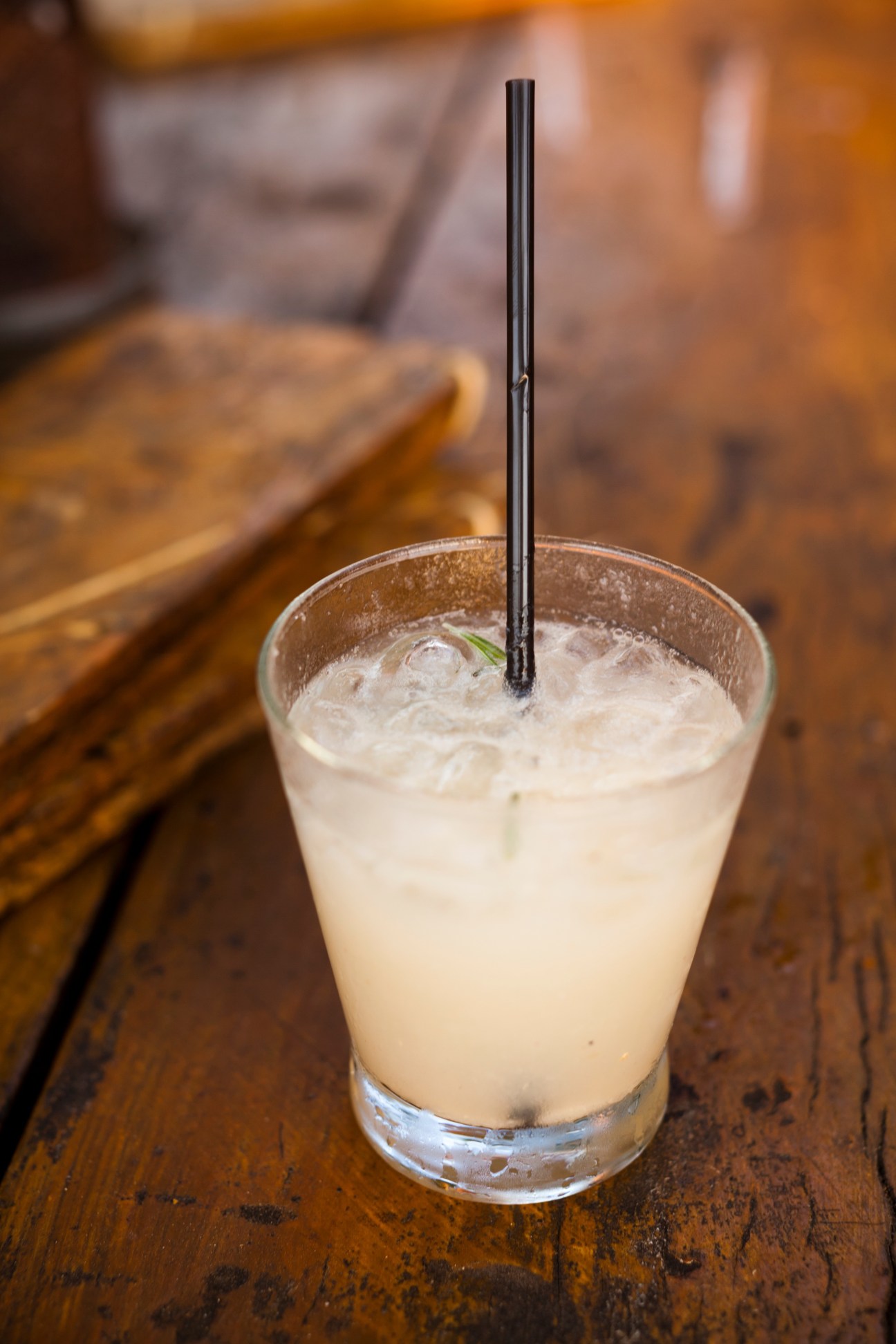 A pale beverage with ice and a black straw in a glass on a wooden surface.