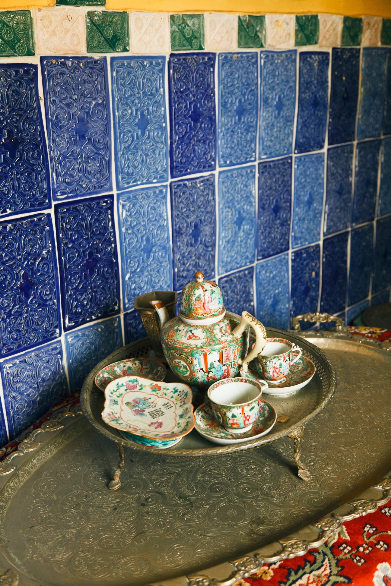 Ornate porcelain tea set with teapot and cups displayed on brass tray against blue zellige tilework.