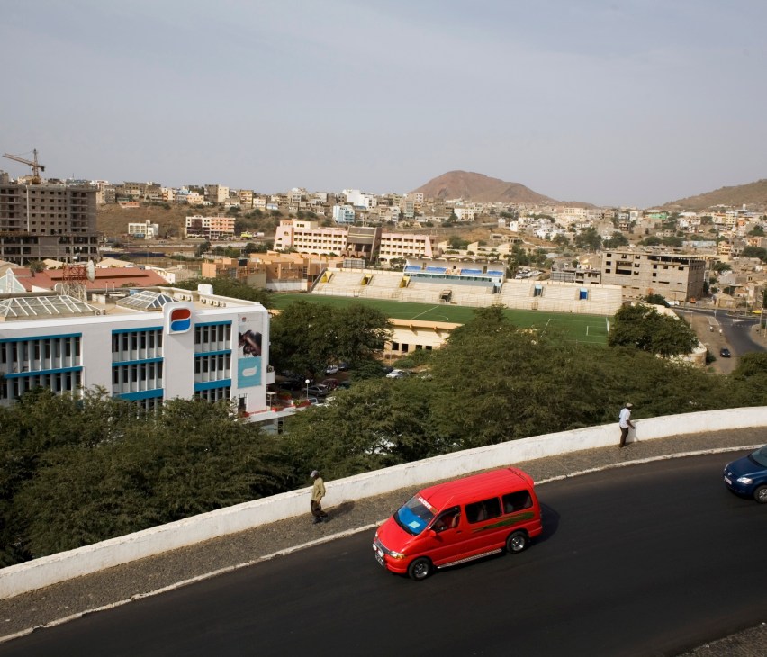 Aerial view of Praia, Cape Verde's capital, showing modern buildings, residential neighborhoods, and arid hillsides with a red vehicle on a new road.