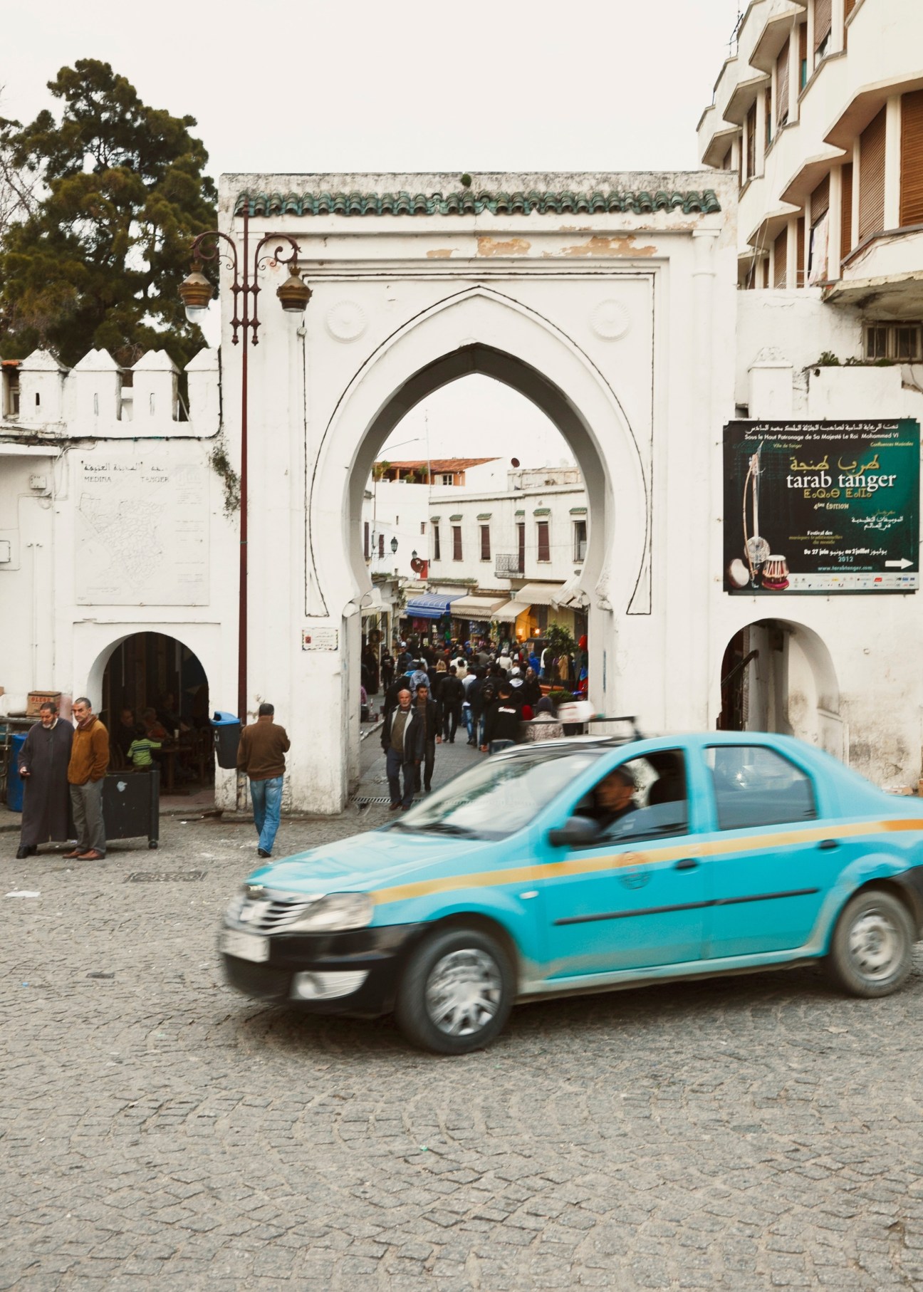 Blue taxi passing through an ornate white archway entrance to the Grand Socco square in Tangier with pedestrians and shops visible beyond.