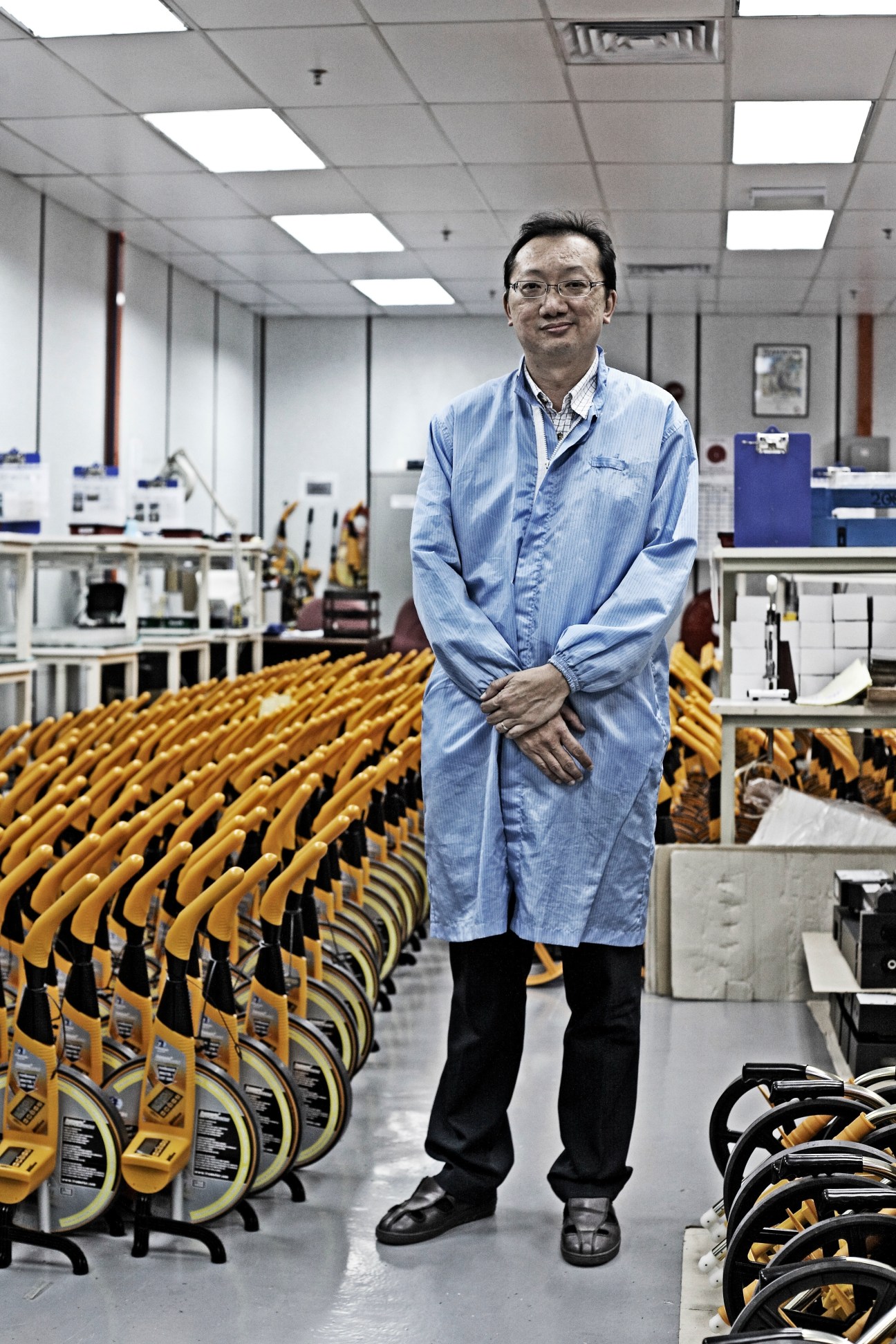 David Lim, manager of a factory making distance-measuring instruments for British firm Trumeter, stands in a factory surrounded by yellow measuring devices.