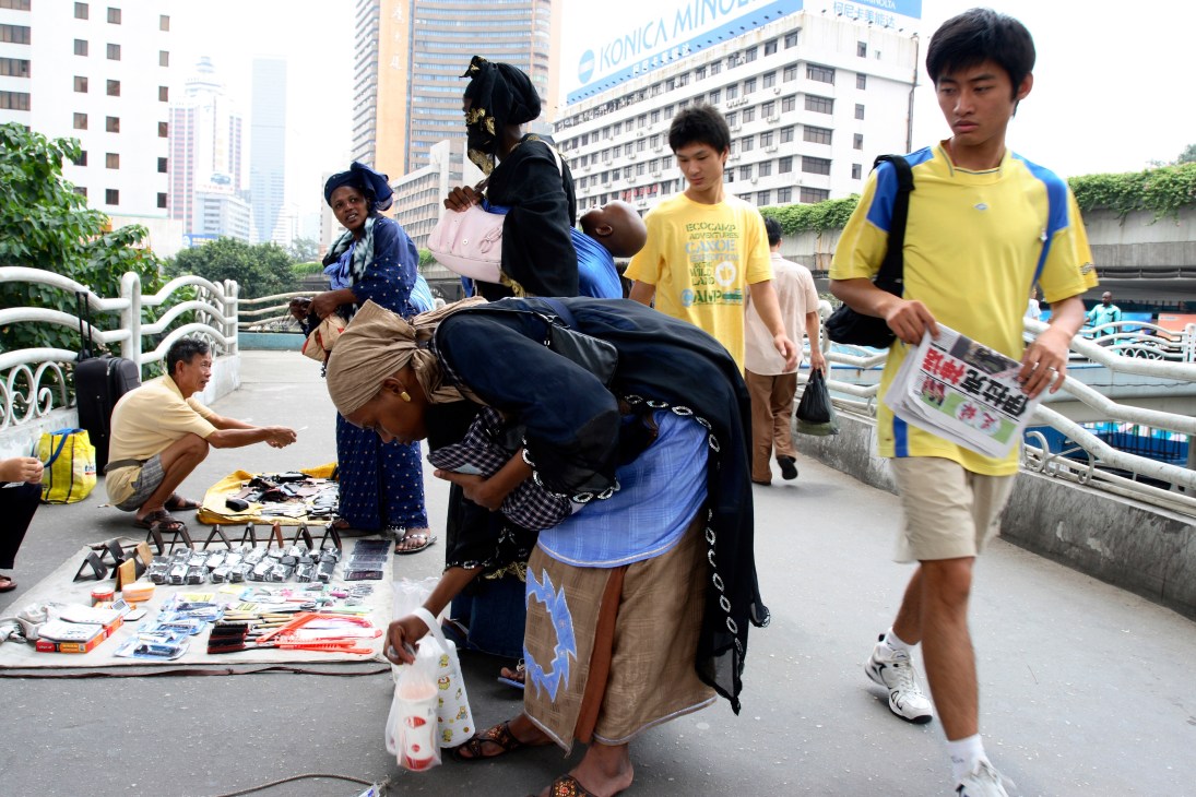 An African woman browses merchandise displayed on the ground by Chinese vendors in an urban plaza with high-rise buildings in the background.