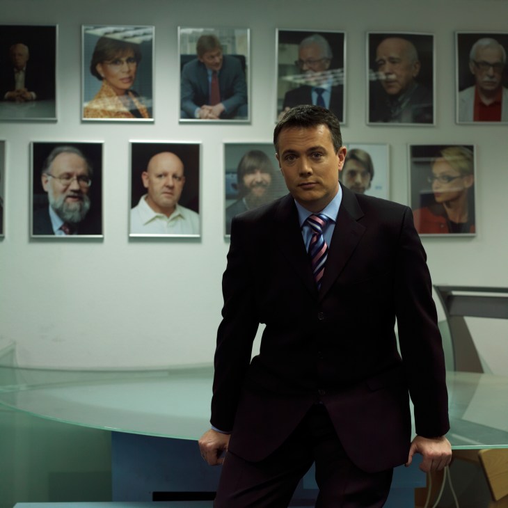 Presenter Kevin Owen stands in a dark suit before a wall of framed portraits of famous guests at Russia Today studios.