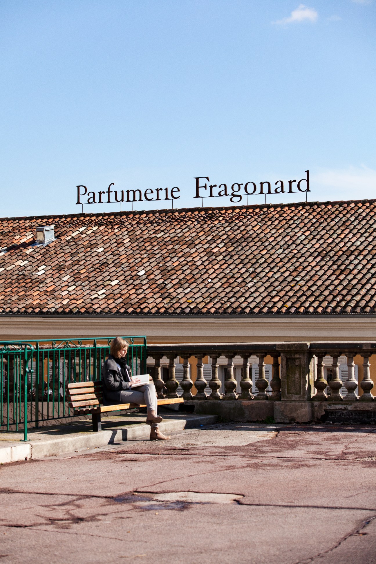 Person sitting on a bench outside Fragonard's historic Grasse factory with terracotta roof and signage.