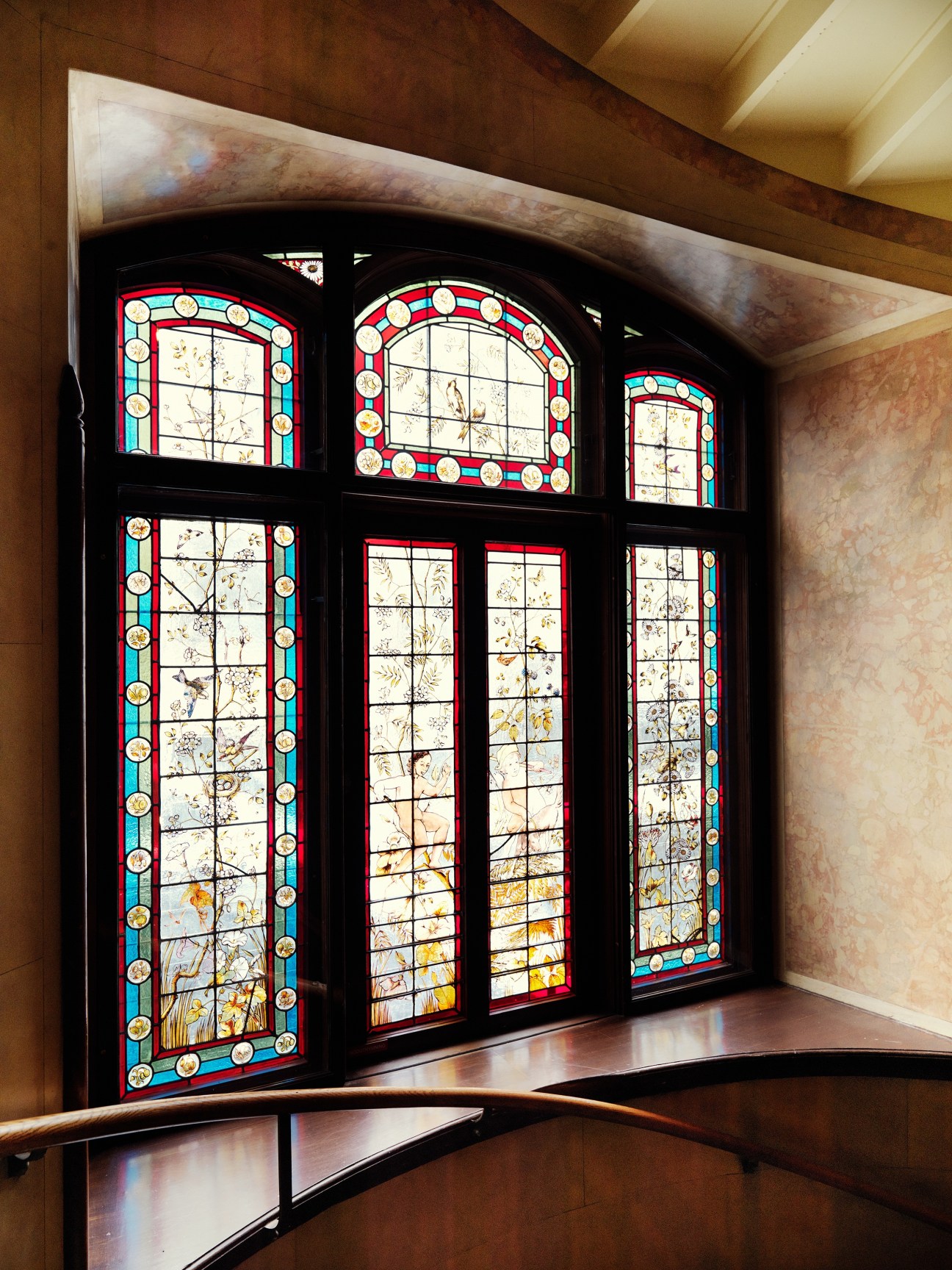 Ornate stained glass windows with red and turquoise borders and decorative circular medallions in a dark wood frame within a stairwell.