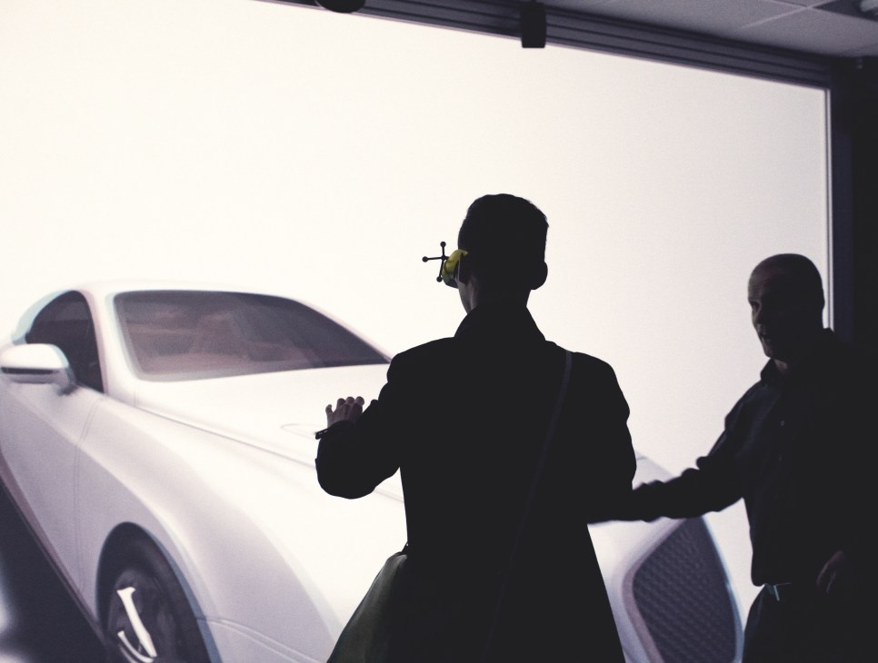 Silhouetted figures examining a white car in a bright showroom or presentation space.