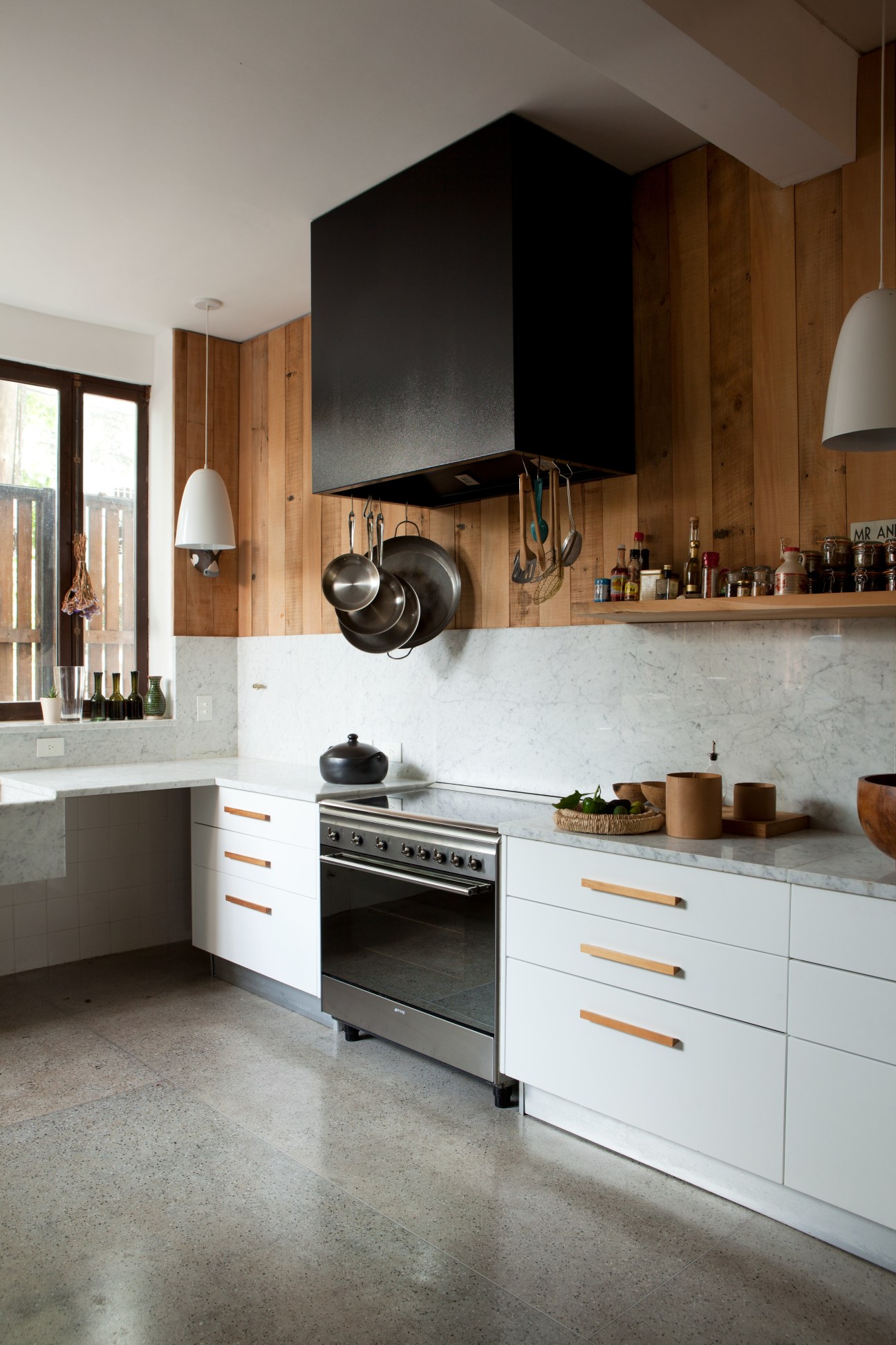 Modern kitchen with wood paneling, marble countertops, black range hood, and hanging cookware.