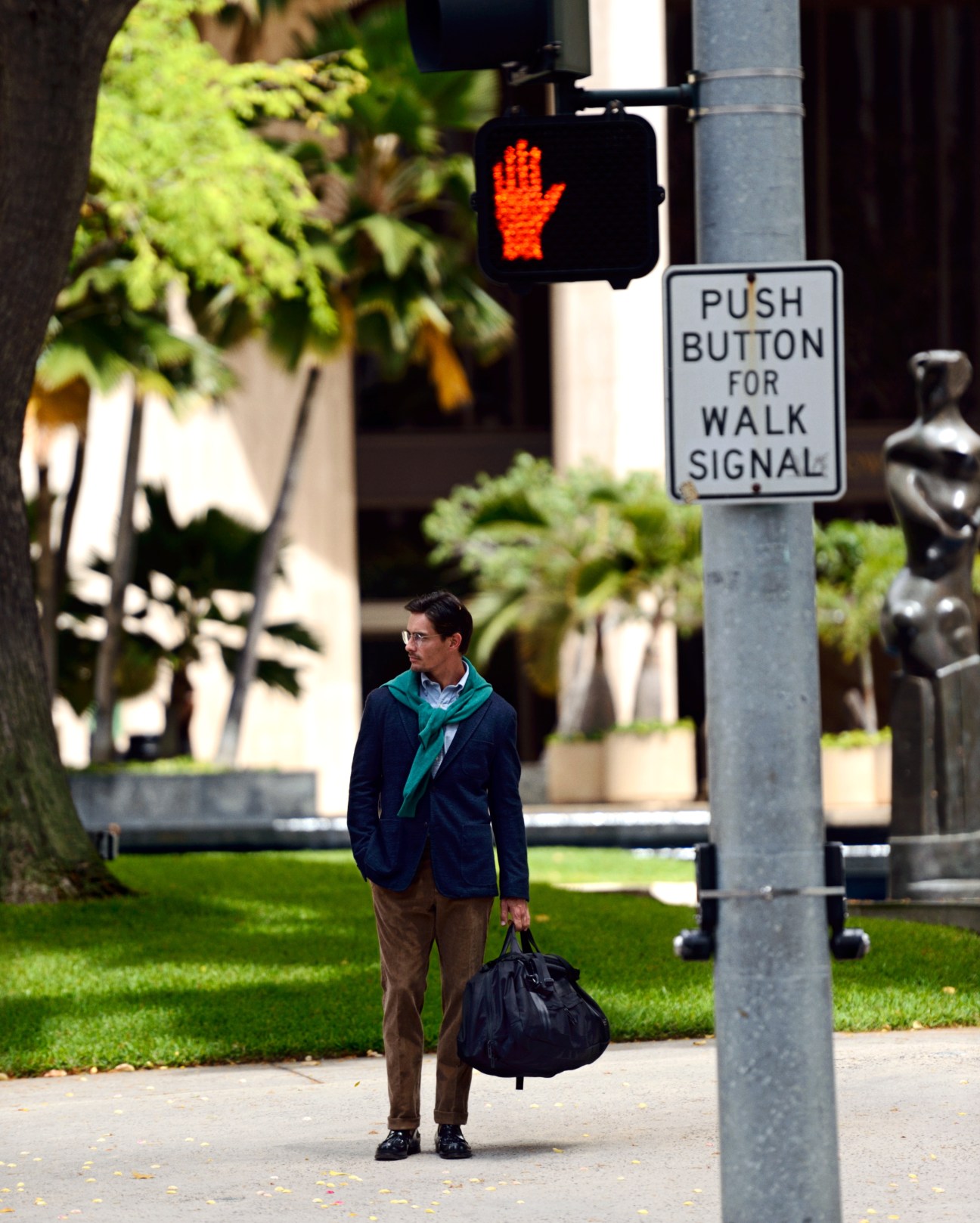Man in navy blazer and brown trousers holding black bag beneath pedestrian walk signal.