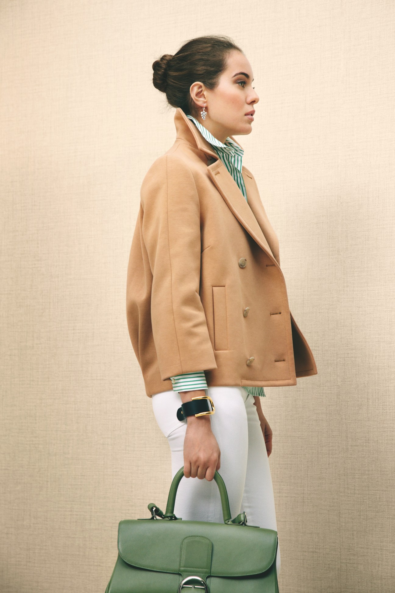 Woman wearing a camel blazer, striped shirt, white trousers, and green handbag with jeweled earrings.