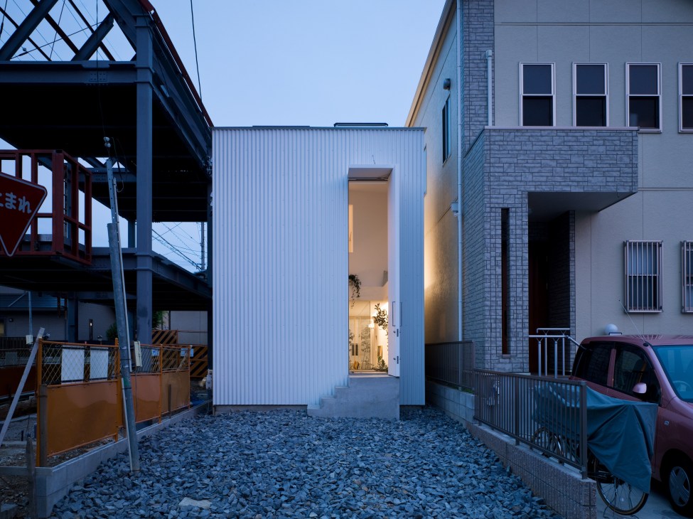 Modern white corrugated metal house exterior with tall narrow entrance door, set in suburban Japanese neighborhood at dusk.
