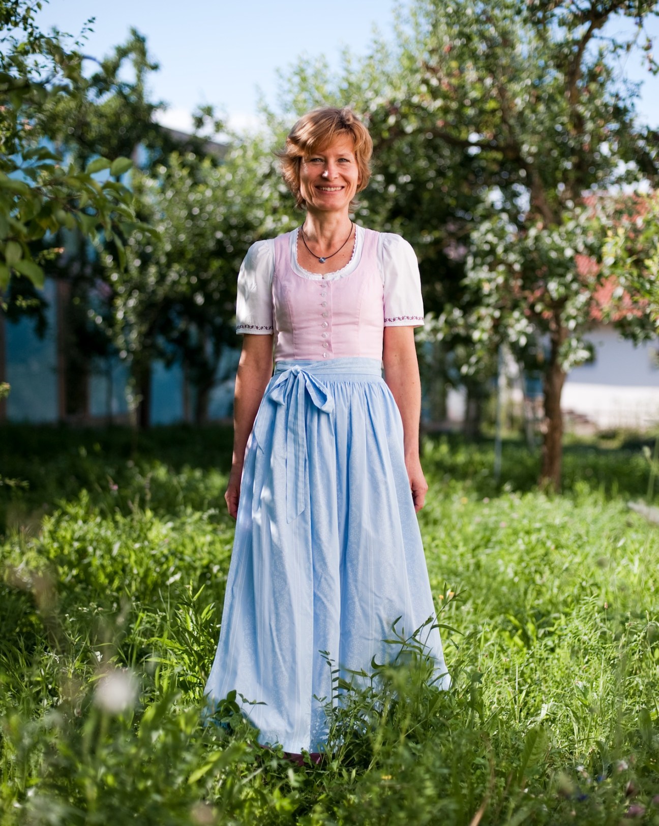 Woman in pink blouse and blue skirt standing in an orchard with fruit trees.