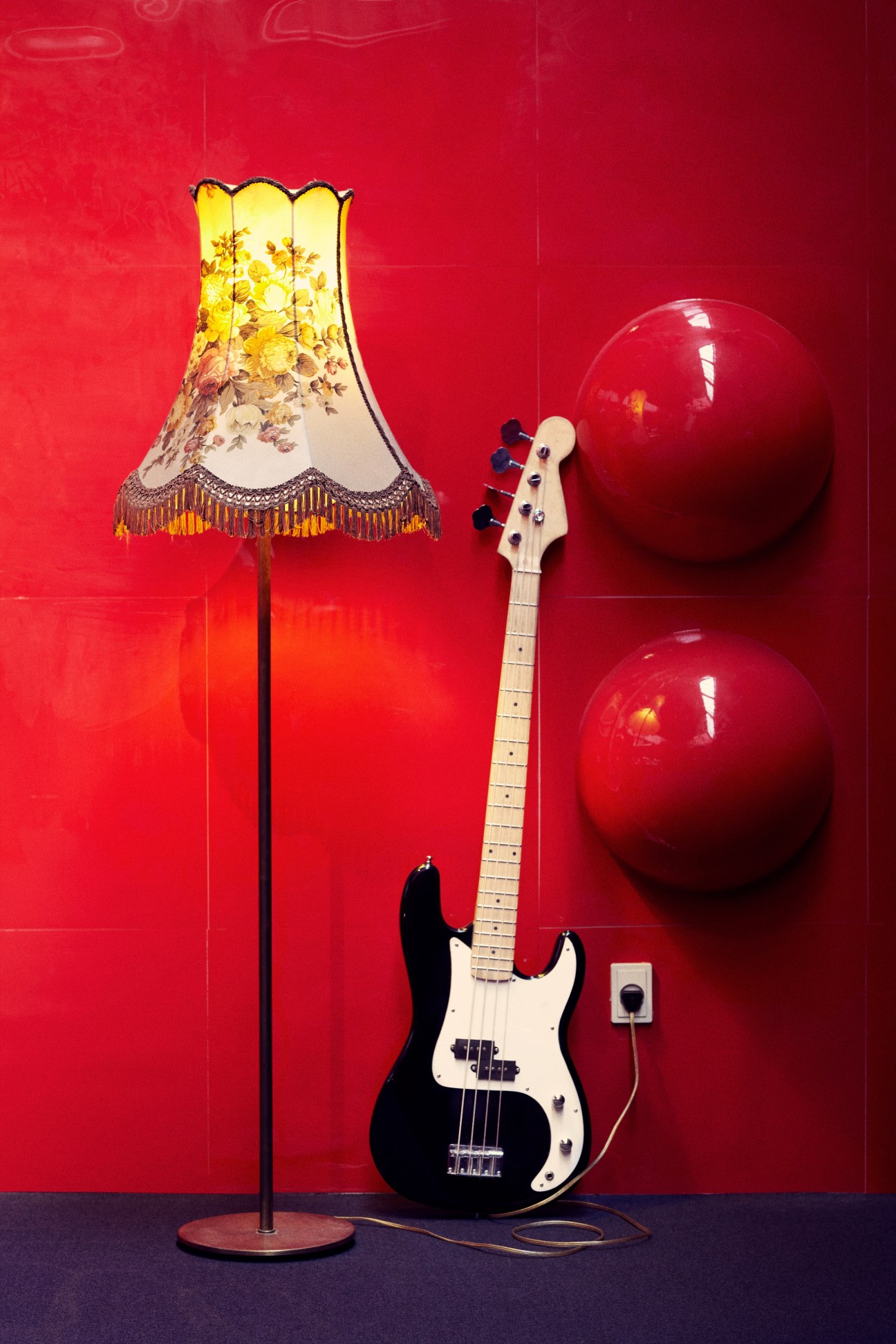 A black electric bass guitar with white pickguard leaning against a red wall next to a decorative lamp and red spheres.