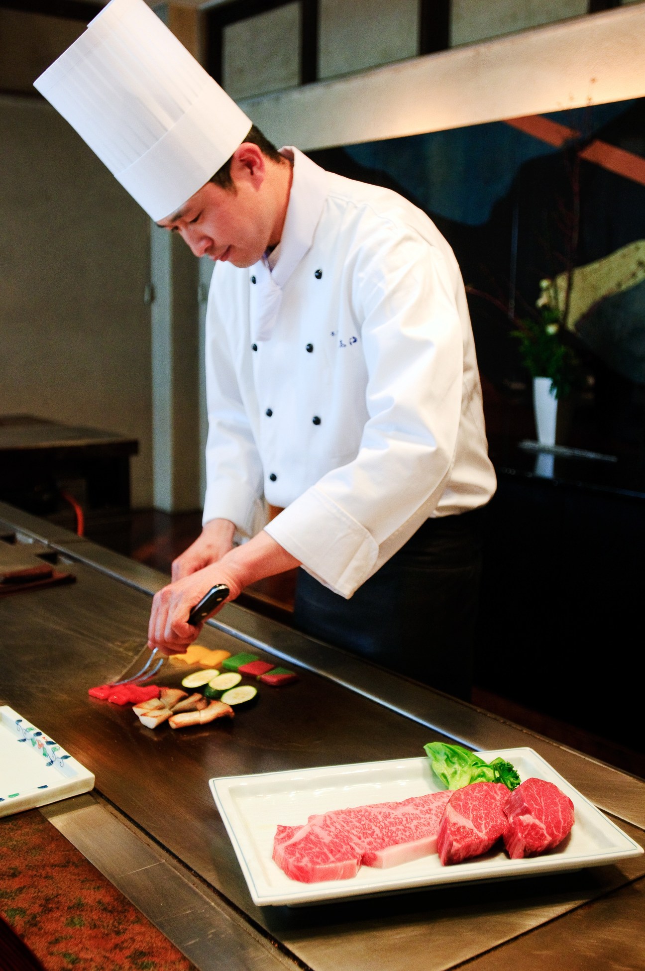 Teppanyaki chef Tomiyuki Kohada in white chef's coat and hat preparing vegetables on a hot steel griddle with marbled Omi beef plated in foreground.