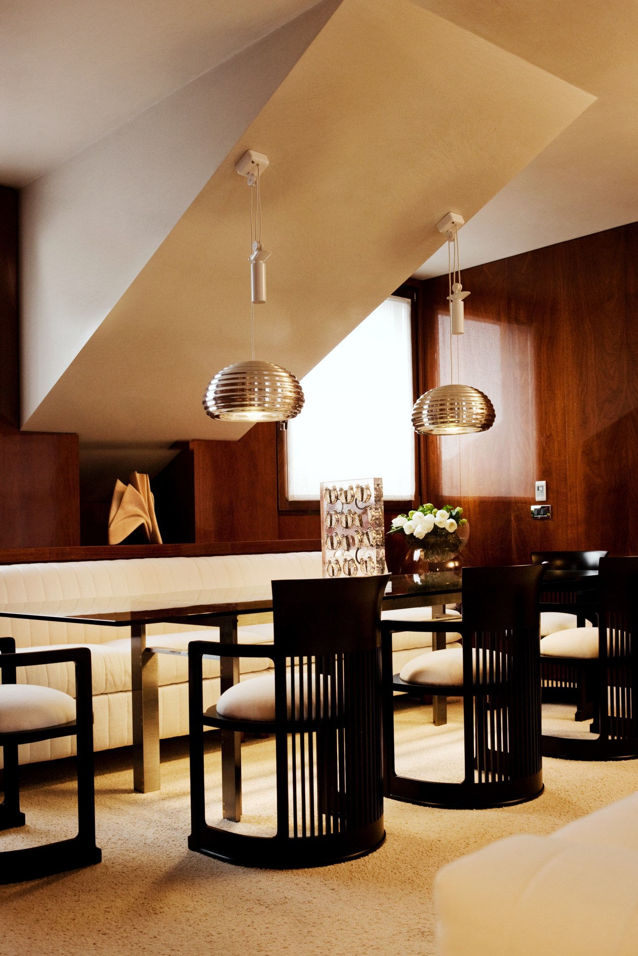 Modern dining room with Carlo Scarpa table, Frank Lloyd Wright chairs, and brass pendant lights under angled ceiling.