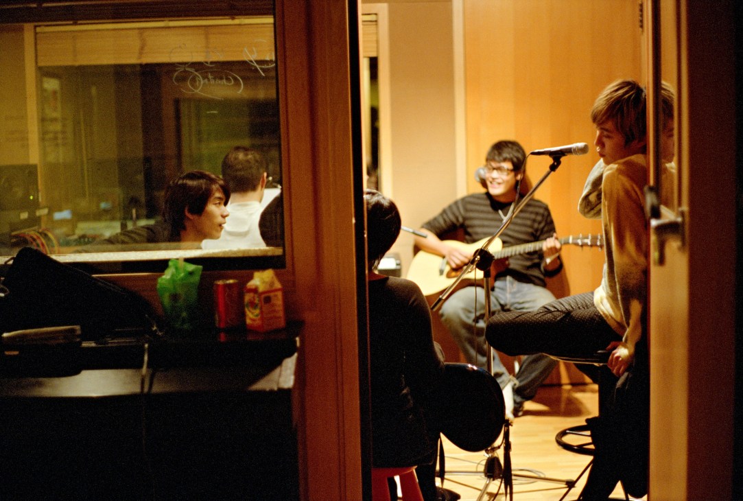 Sodagreen band members performing in a rehearsal room with acoustic guitar and microphone.