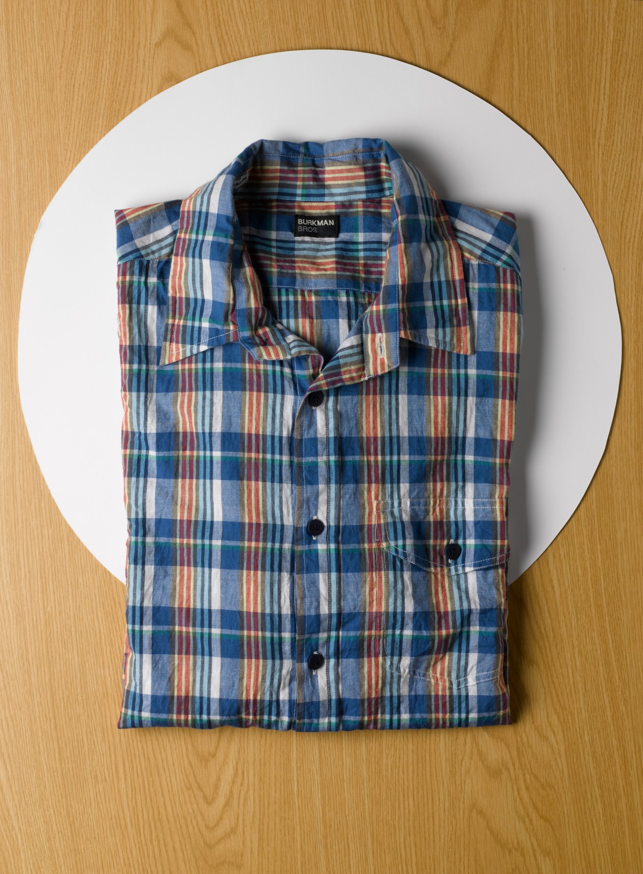 Burkman Bros plaid shirt in blue, red, and white folded on white circle on wooden surface.