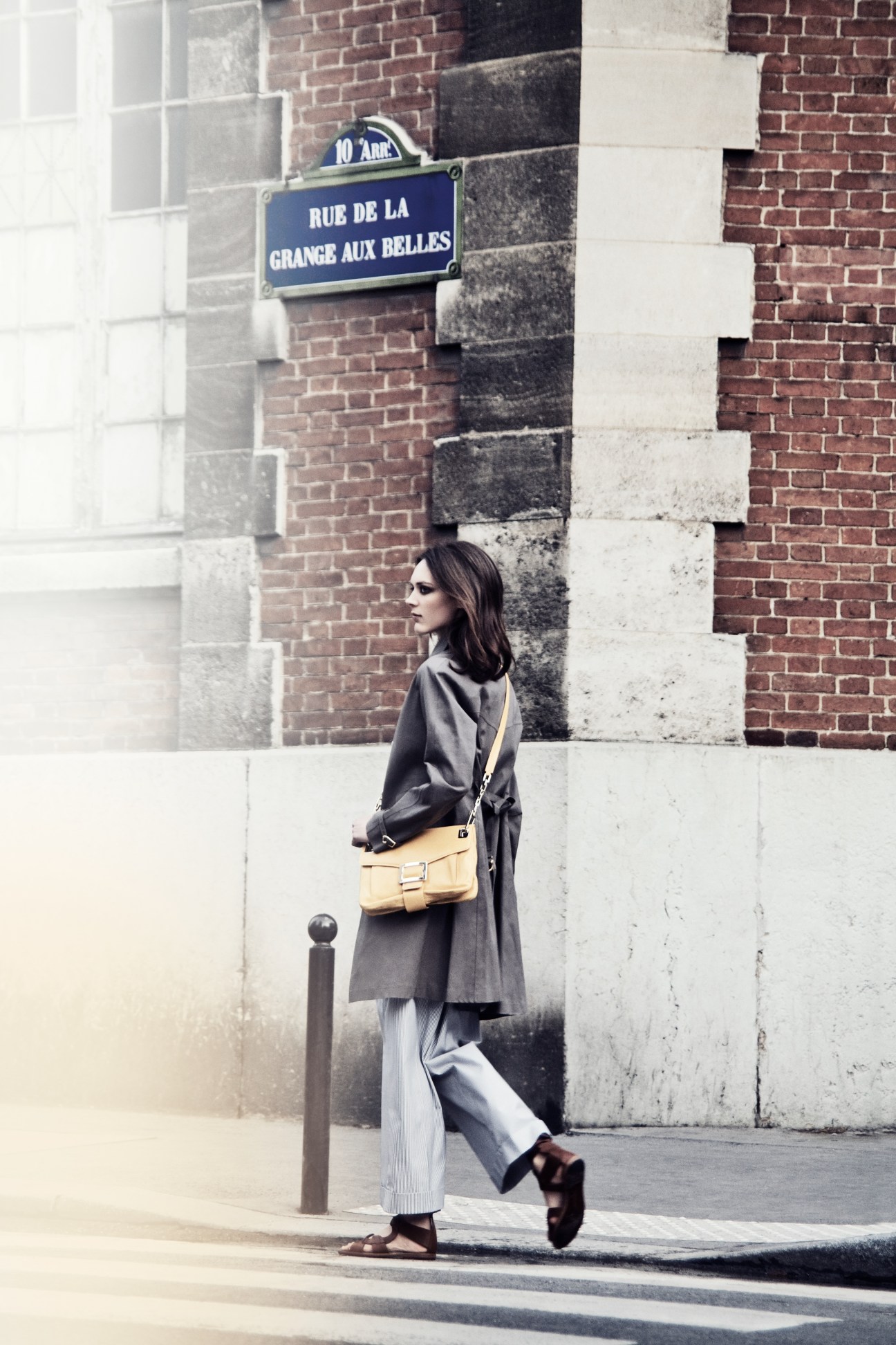 Model in Mackintosh coat and Incotex trousers with Roger Vivier bag stands on Parisian street by brick wall.