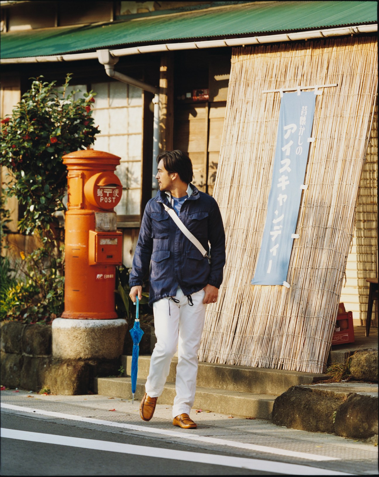 Man in navy jacket and white trousers holding blue umbrella on a Japanese street with red mailbox