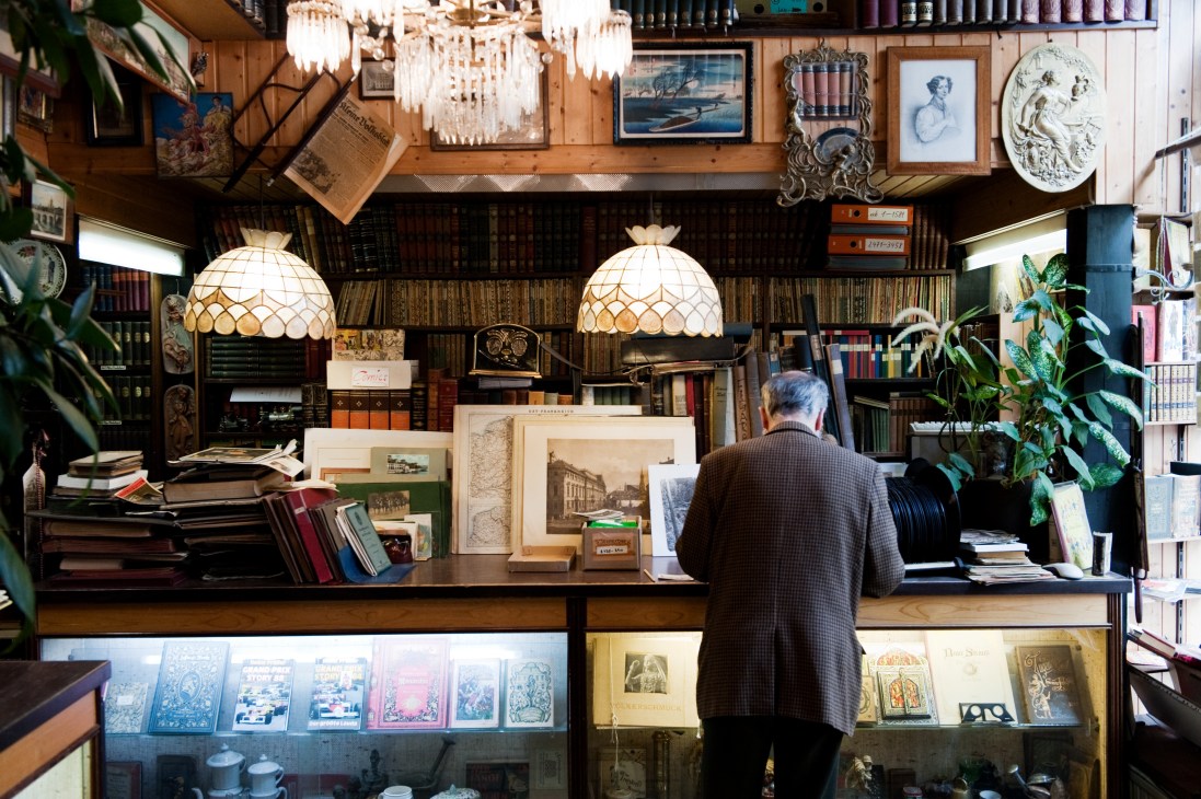 Interior of Bücher-Ernst antiquarian bookshop with ornate ceiling lamps, walls lined with books, and a customer at the counter.