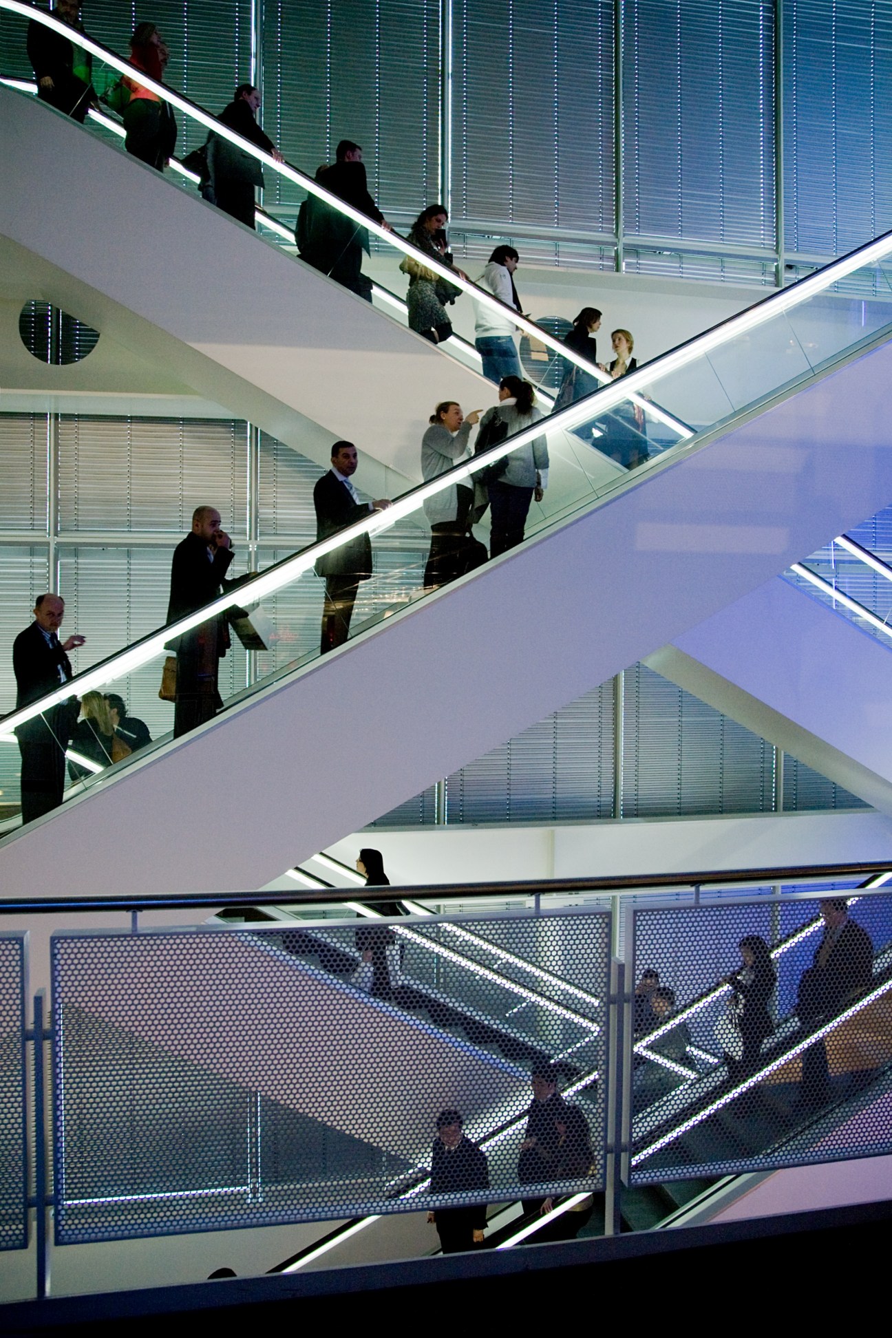 People on multiple escalators inside a modern convention center with glass railings and bright lighting.