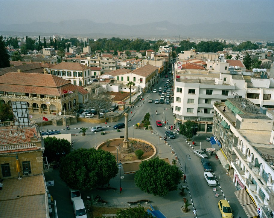 Aerial view of Nicosia showing residential buildings, streets, Turkish flags, and mountains in the distance.