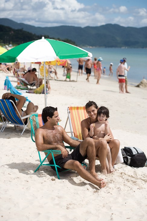 A family relaxes on a sandy beach under a green and white umbrella with mountains and water visible in the background.