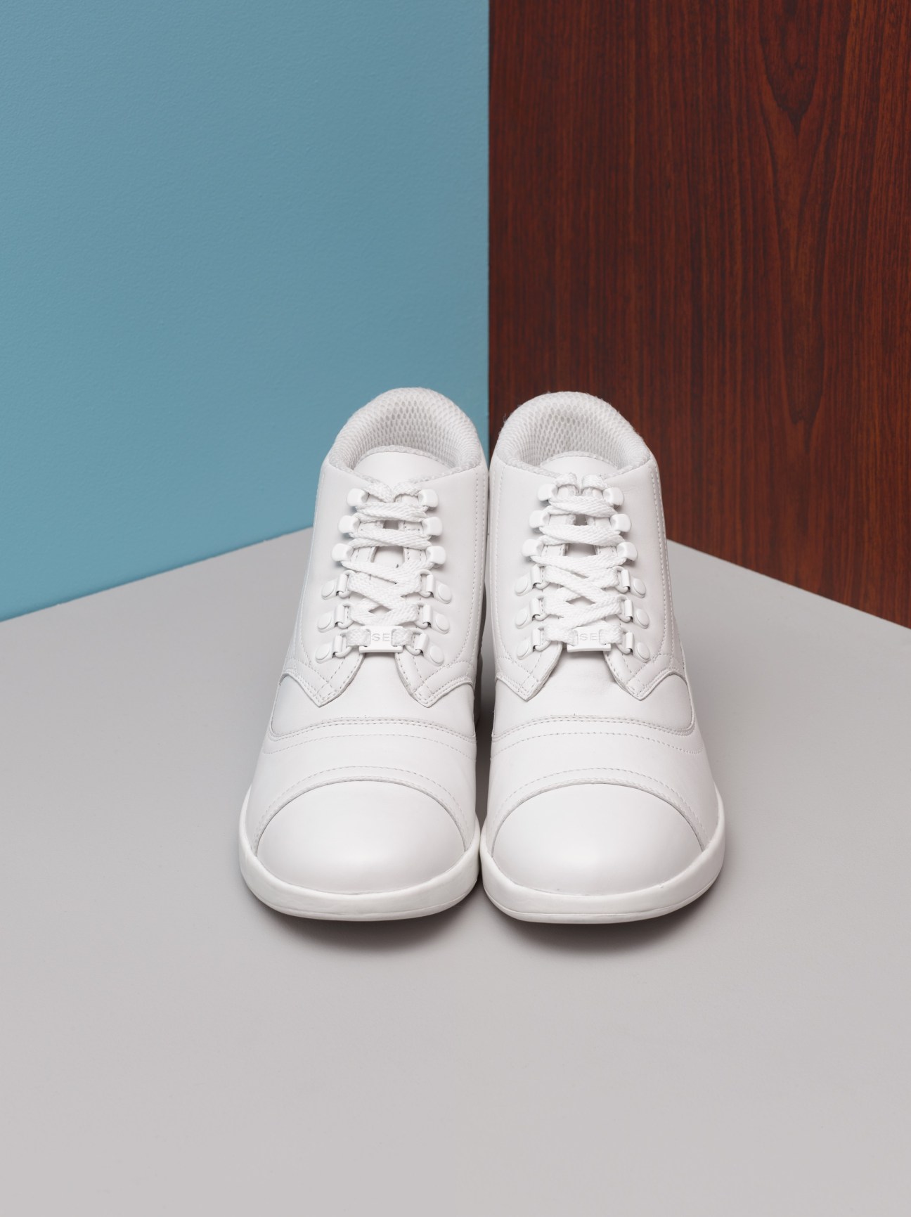 White leather Gram trainers displayed against a blue and wood-grain background.