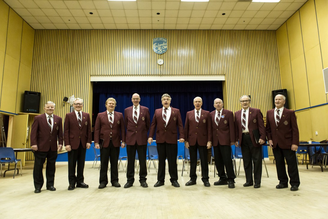 Nine men in matching maroon blazers with emblems stand in a line on a wooden stage in a yellow hall.