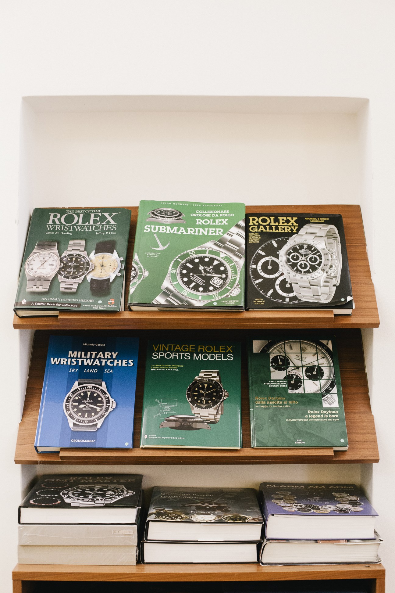 Wooden wall shelves displaying coffee table books about luxury watches including Rolex, Submariner, and Palex models.