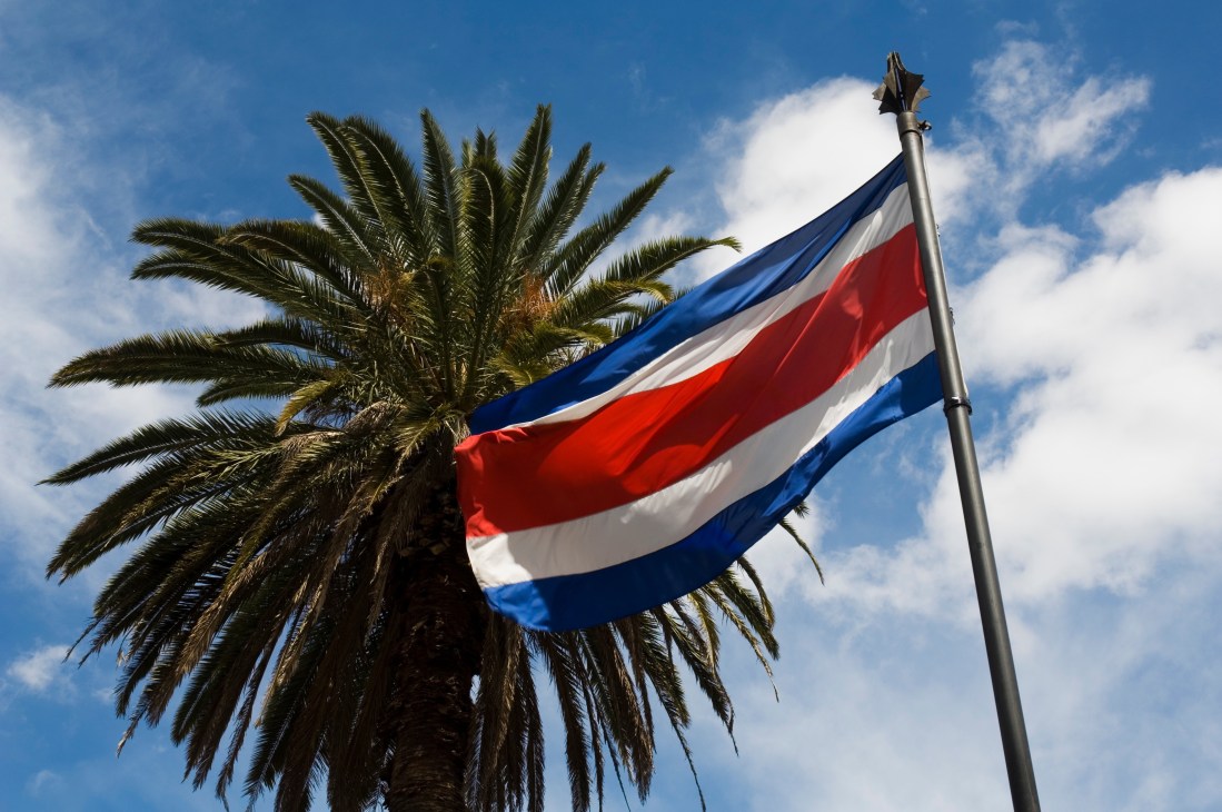 Costa Rican flag waving on a pole beside a palm tree against blue sky with white clouds.