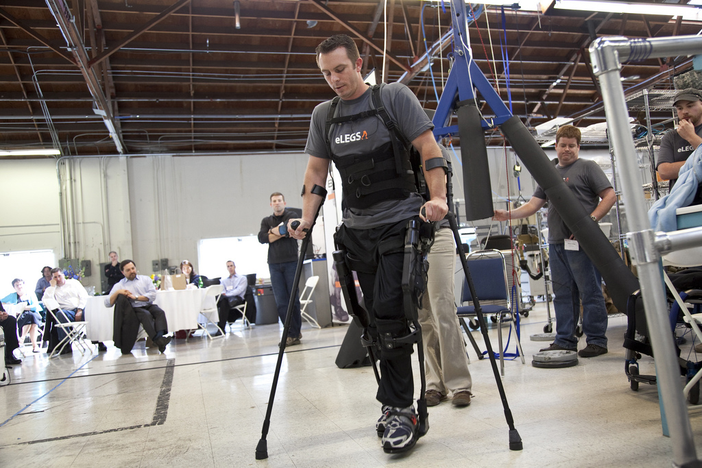 Person wearing Ekso Bionics exoskeleton demonstrates walking at demonstration event with audience seated in background.