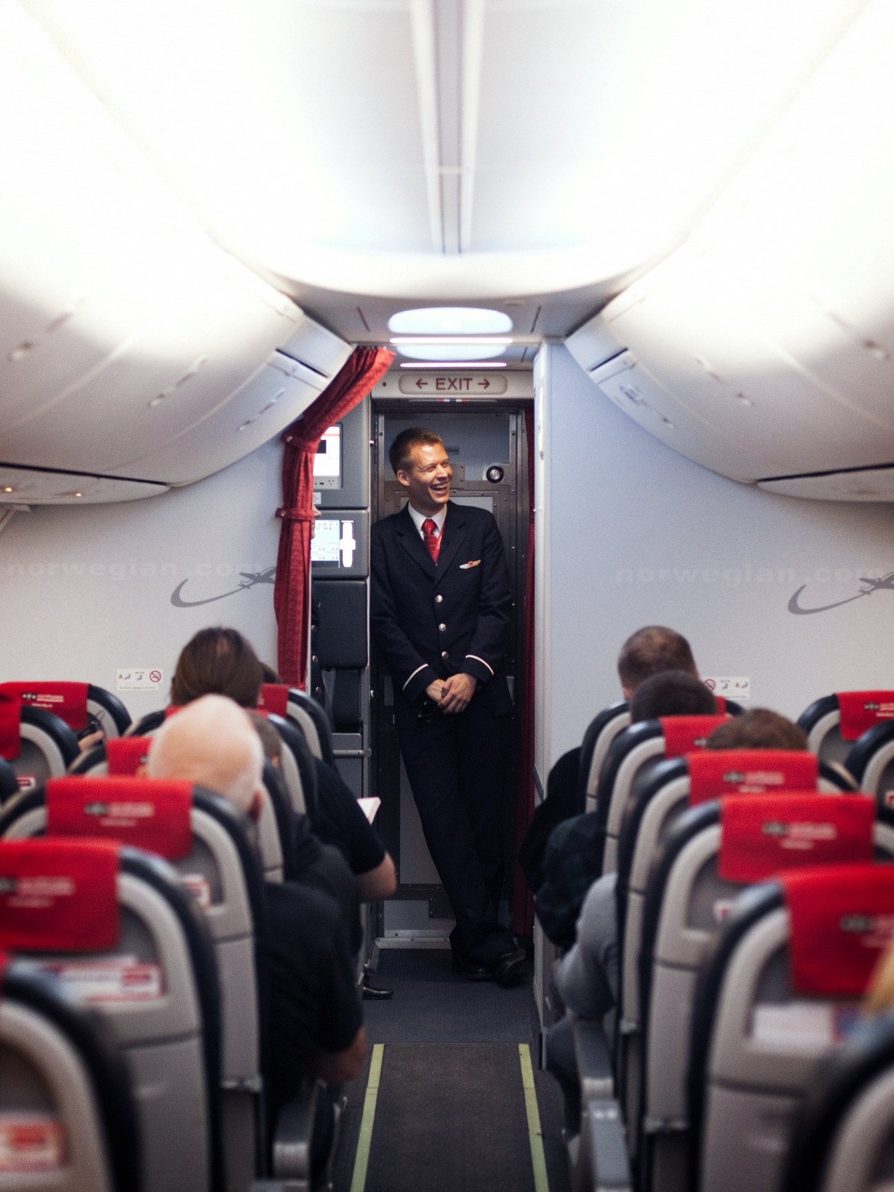 Flight attendant in uniform standing in aircraft cabin aisle with red seat headrests visible.