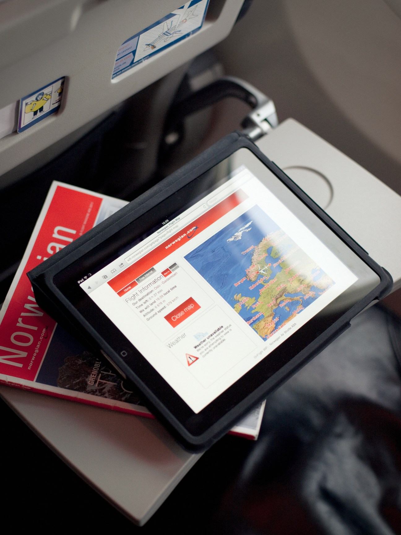 Tablet displaying Norwegian airline website in aircraft seat pocket with safety information visible overhead.