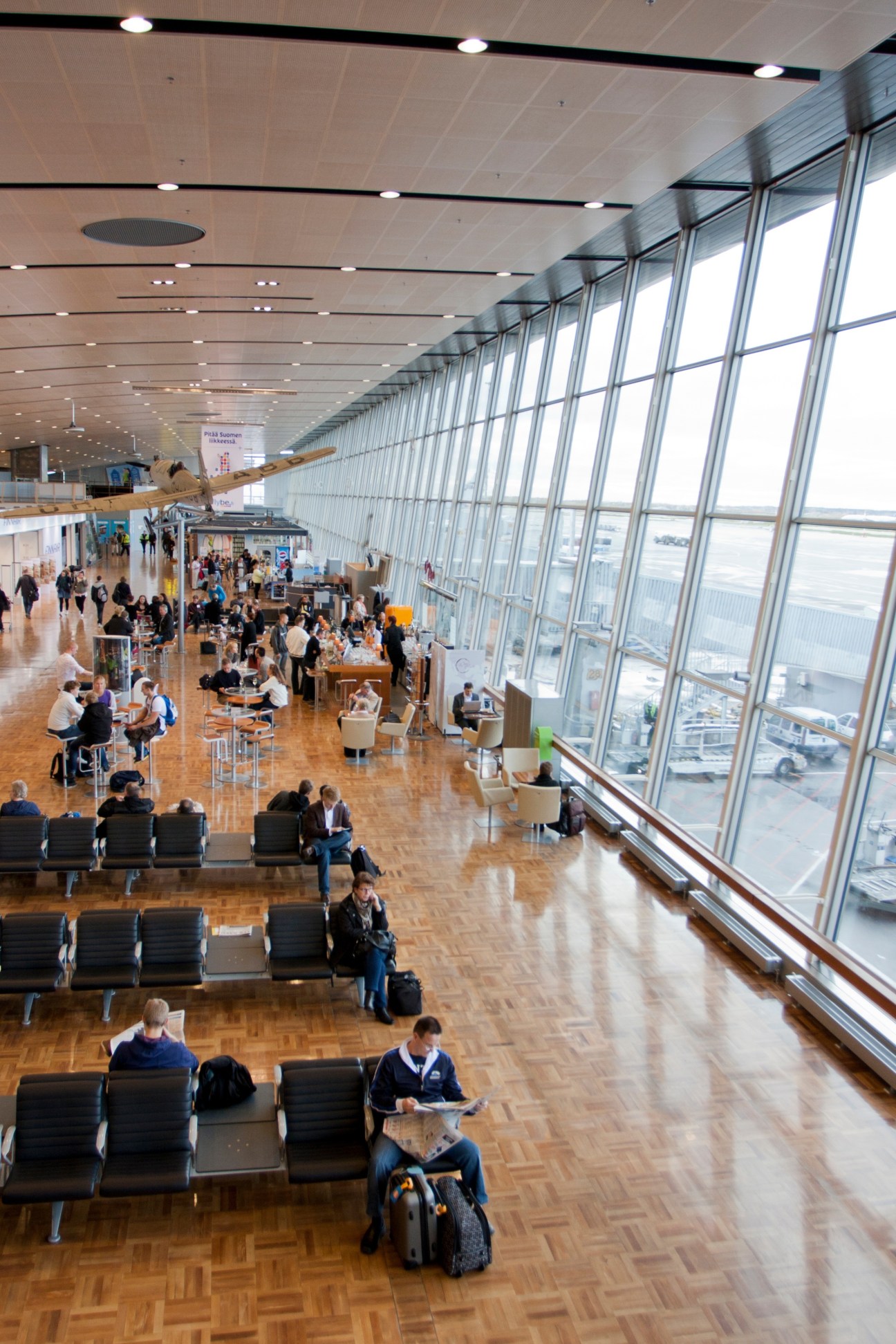 Interior view of a modern airport terminal with wooden flooring, floor-to-ceiling windows, seating areas, and travelers.