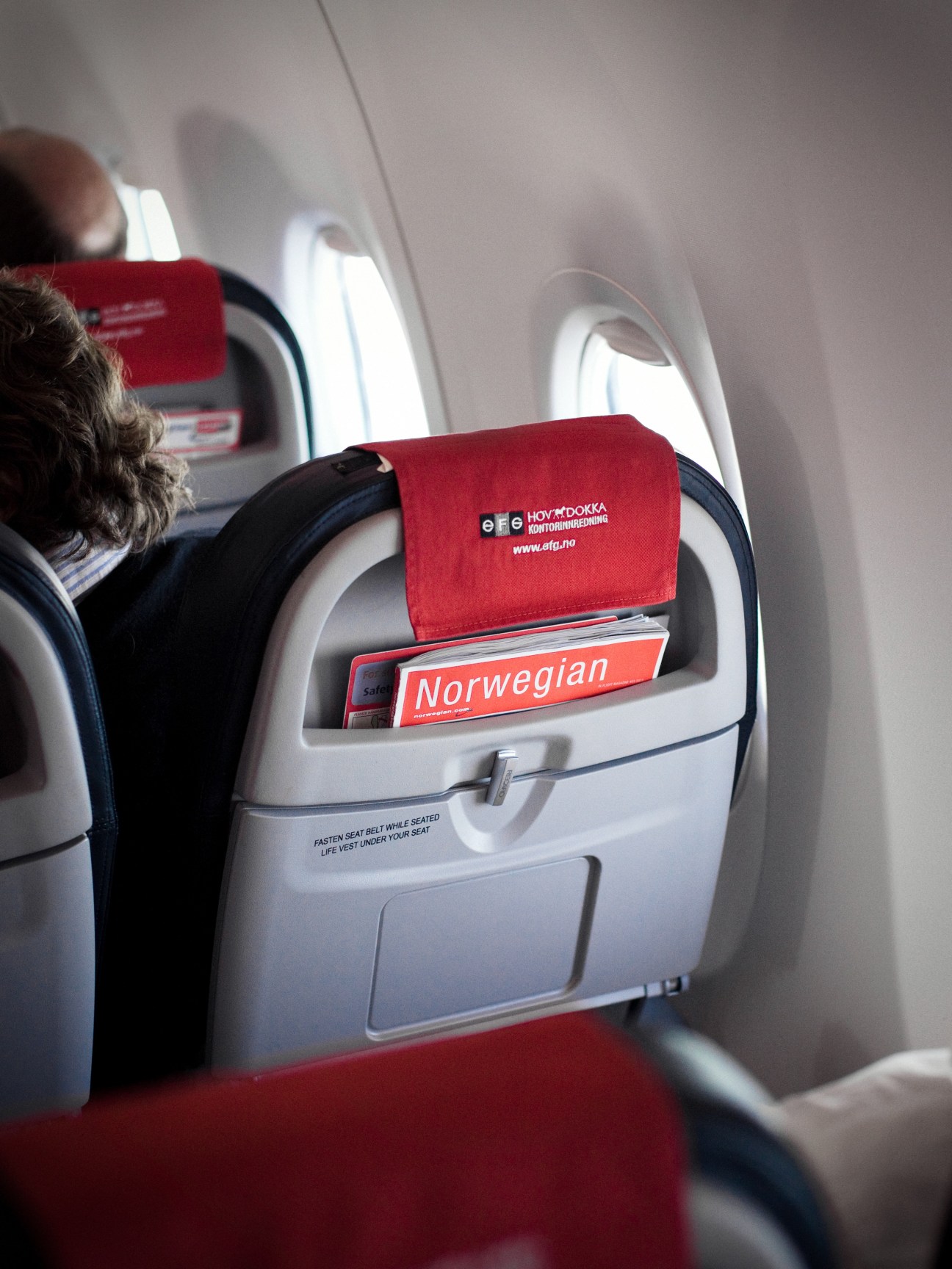 Norwegian airline seat with red headrest cover and amenity kit in aircraft cabin.