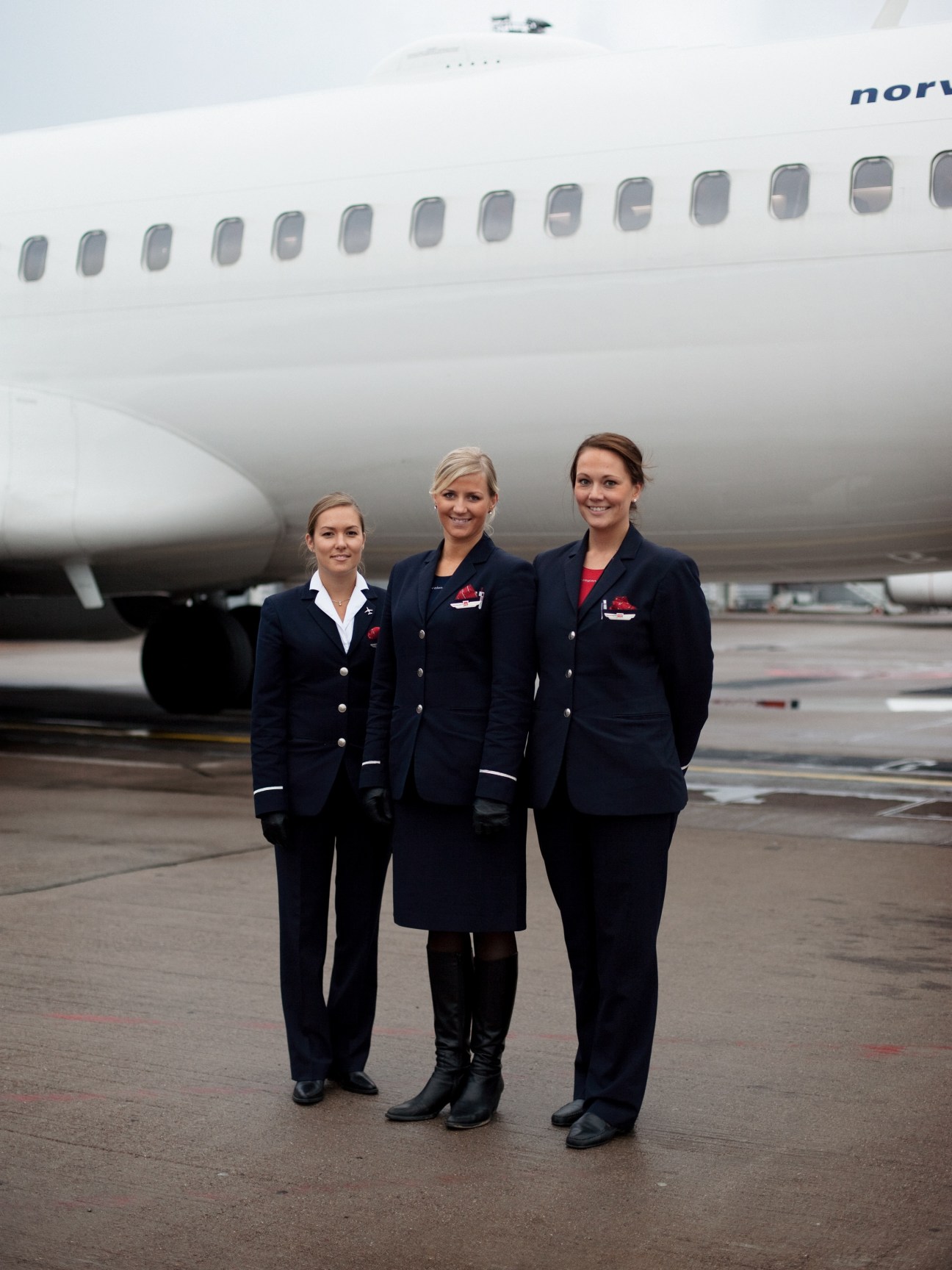Three Norwegian airline flight attendants in navy uniforms standing in front of a white aircraft on tarmac.