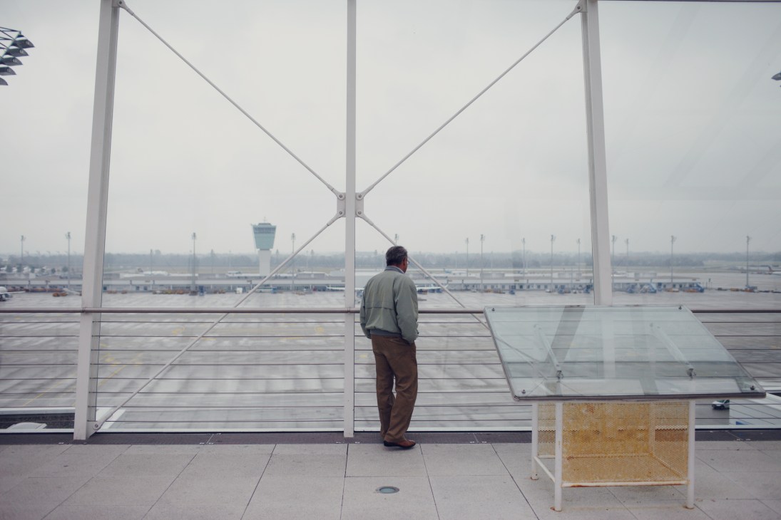 Man viewing airport tarmac and control tower through large windows at observation deck.