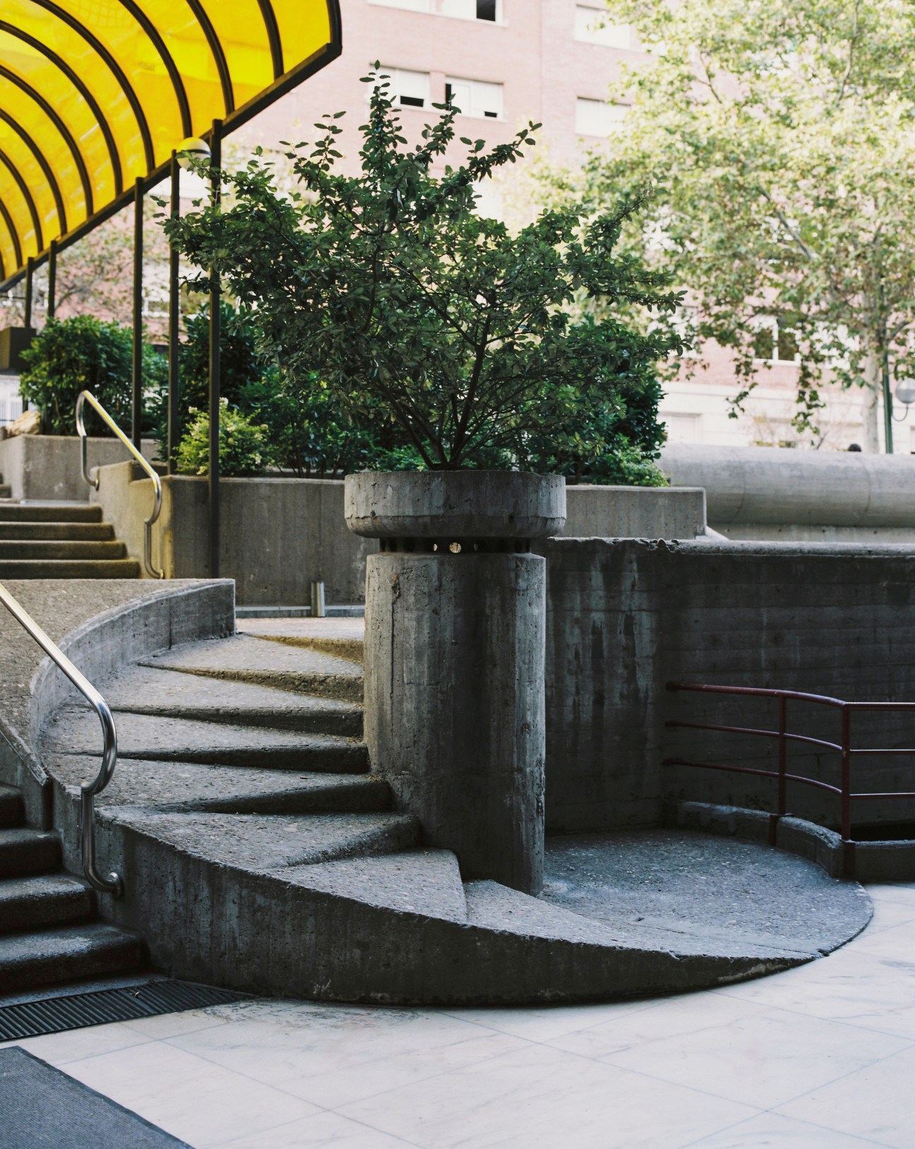 Concrete street-level entrance with yellow canopy, stairs, and planted trees at Torres Blancas.