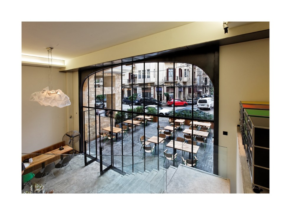 Modern interior space with cream walls, large black-framed glass doors opening to a street-level café with wooden tables and chairs.