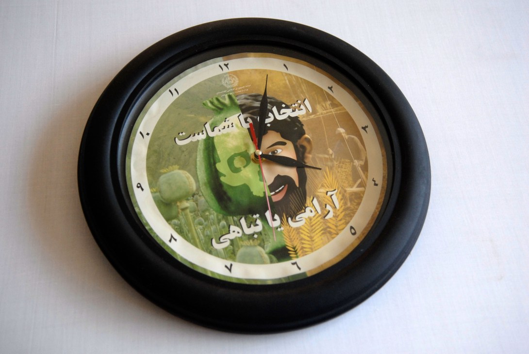 Wall clock with black frame featuring an anti-opium message in Dari or Pashtu text and illustrated imagery.