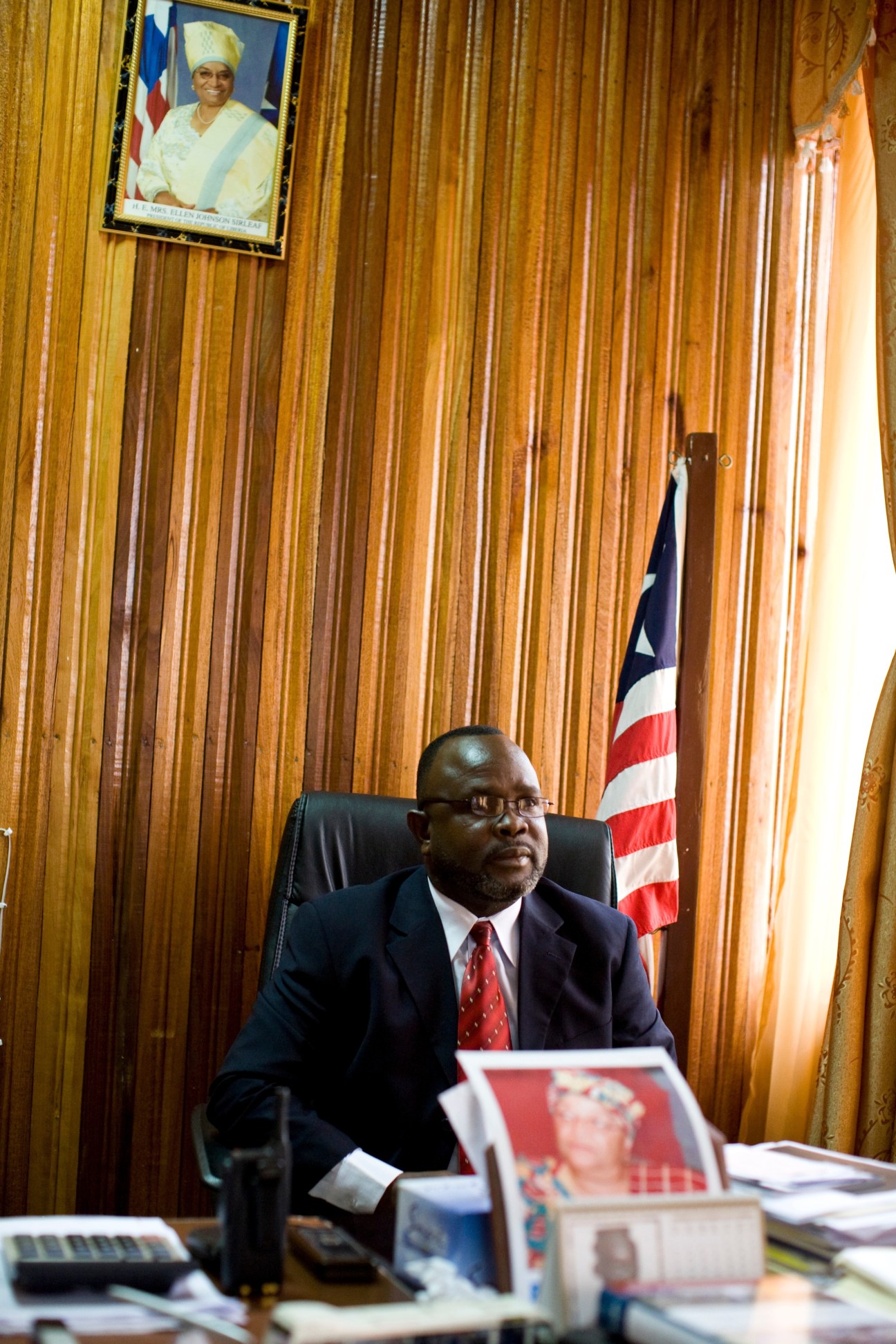 Transport minister Alphonso Gaye sits at desk in wood-paneled office with Liberian and American flags.