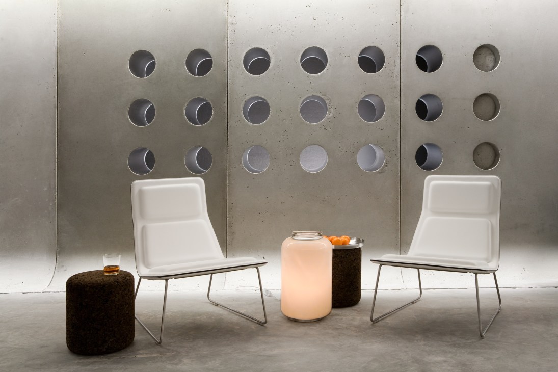 Modern bar lounge with white chairs, concrete wall, circular perforations, and glowing pendant lights.