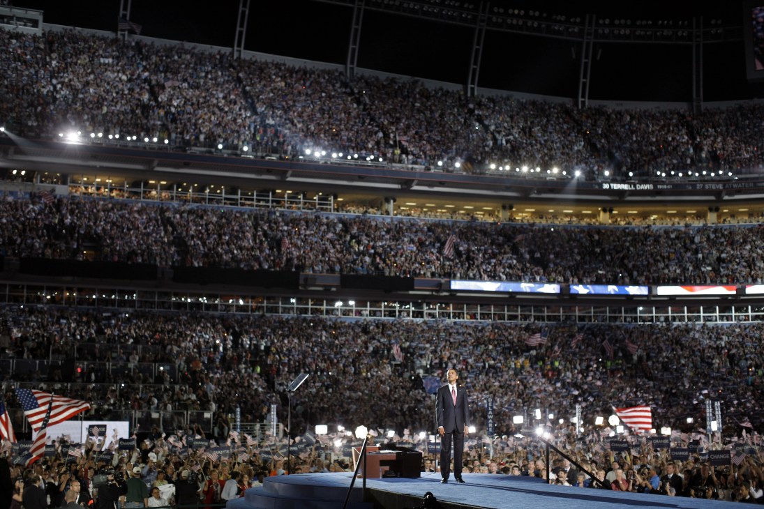 Politician speaking at podium in large stadium filled with crowds and American flags.